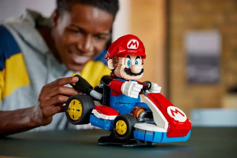 A large Mario Kart Lego set on a table seen side on. A person touches the back of the kart and smiles with glee.